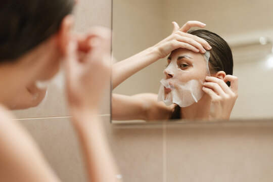 Young woman applying facial mask in front of mirror, showcasing skincare routine and self-care moments with a focus on beauty and wellness.