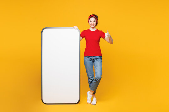 Full body young happy smiling woman wear red t-shirt casual clothes big huge blank screen mobile cell phone smartphone with area show thumb up isolated on plain yellow background. Lifestyle concept. - Powered by Adobe