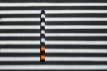 A cigarette with an orange filter lies on a white windowsill in the shadow of blinds that resemble...