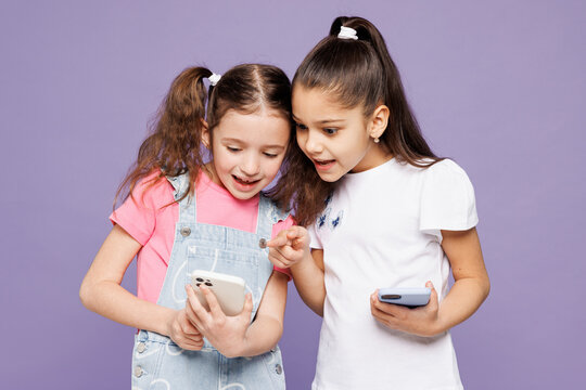Little smiling fun two friends kids children girls 9-10 years old wear casual clothes together hold in hand use show look at mobile cell phone isolated on plain purple background. Lifestyle concept.