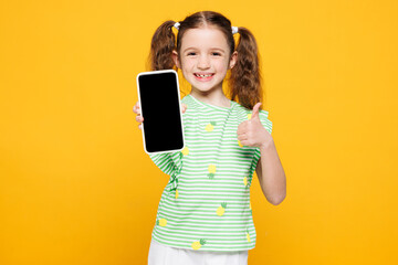 Fototapeta premium Little kid child girl 8-9 years old wears green t-shirt hold in hand use mobile cell phone with blank screen area show thumb up isolated on plain yellow background. Mother's Day love family concept.