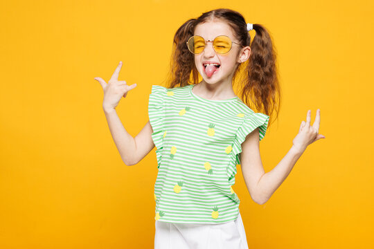 Little smiling cheery cool kid child girl 8-9 years old wears green t-shirt sunglasses do horns up gesture show tongue isolated on plain yellow background. Mother's Day love family lifestyle concept.