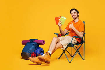 Young happy traveler man sit in chair backpack with stuff mat hold snack isolated on plain yellow...