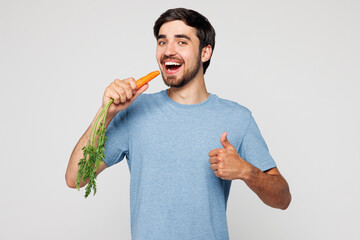 Young smiling happy man wear blue t-shirt casual clothes hold in hand eat bite raw carrot show...