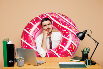 Young bored employee business man wear white shirt tie pink donut rubber ring sit work at office...