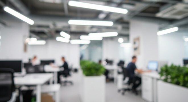 Blurred view of a modern, bright office space with employees working at desks, illuminated by overhead lighting.