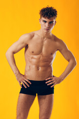 Fototapeta premium Confident young man with curly hair posing in black swimwear against bright yellow background, showcasing fitness and health awareness.