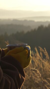 Vertical video of girl in coat and yellow gloves holding tea mug against backdrop of mountains and meadow on mountain slope during autumn, captured in video footage