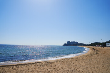 Fototapeta premium Sunny coastal beach with gentle waves, clear blue sky, and distant buildings along the shoreline