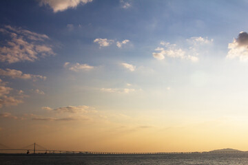 Scenic view of ocean and distant bridge at sunset with warm golden sky and scattered clouds
