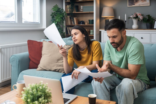 Worried young couple sitting on sofa at home, analyzing financial documents and statistics on laptop and paper, stressed about unexpected expenses, debt, or negative budget report results.