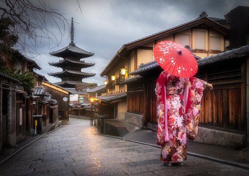 View of a woman in a vibrant kimono and red umbrella walks along a wet, cobblestone street towards a traditional pagoda, Kyoto, Kyoto, Japan. - Powered by Adobe