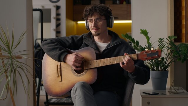Smiling Caucasian musician guy playing guitar solo at home hipster man in headphones male hobby relaxing music performance song happy artist enjoying play acoustic musical instrument practicing skills