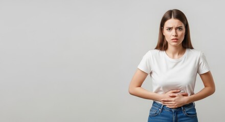 Obraz premium Young woman in casual attire holding her stomach, expressing discomfort in a neutral background