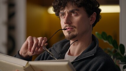 Puzzled pensive Hispanic guy contemplating reading book at home learner read book confused hipster...