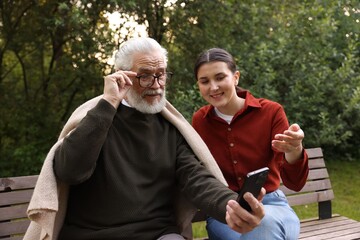 Smiling caregiver and elderly man in glasses using smartphone on bench outdoors