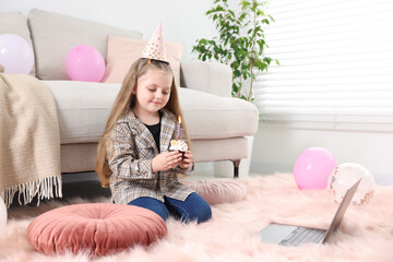 Birthday celebration. Little girl with cupcake having video call via laptop at home