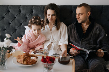 
A joyful child, with curlers in her hair, shares a cozy morning in bed with adults, as they read...