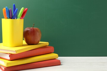 Different school stationery and red apple on white wooden table against blackboard, space for text