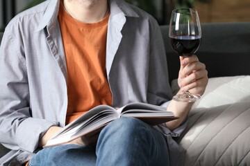Woman with glass of wine reading book on sofa at home, closeup