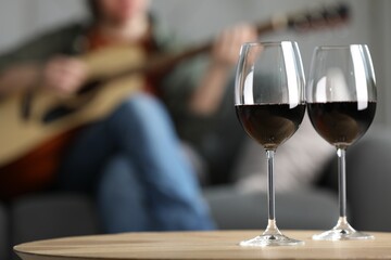 Glasses with red wine on table and man playing guitar indoors, selective focus