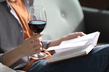 Woman with glass of wine reading book on sofa at home, closeup