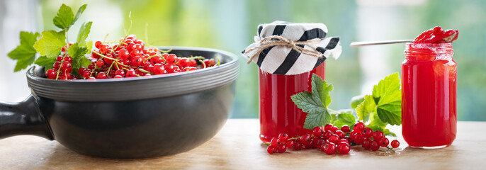 Fresh homemade red currant jam in jar with fresh picked currants in a bowl on wooden table against light background. Horizontal close-up. 