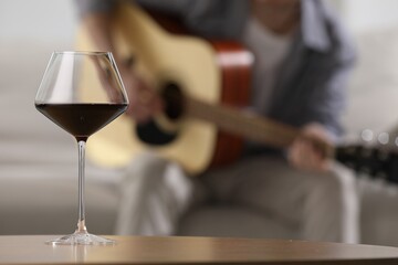 Glass with red wine on table and man playing guitar indoors, selective focus