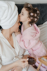 A joyful child in a pink bathrobe with curlers in her hair, laughs happily while looking at her...