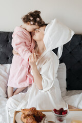 A loving moment between an adult woman and a child in bathrobes, facing each other on a bed, with a...