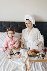 A woman with a towel-wrapped head and a child with curlers read magazines in bed, enjoying...