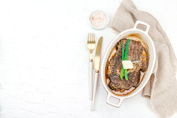 Raw ribeye steak in ceramic baking dish with herbs, spices, cutlery and napkin on white plaster