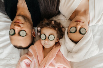 A family enjoys a spa day at home, with cucumber slices on their eyes, relaxing together on a white bed.