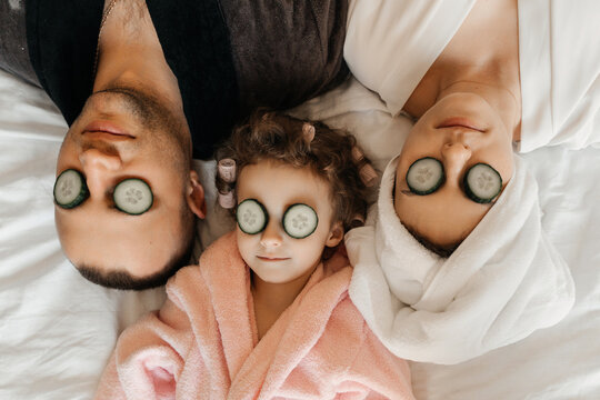 A family enjoys a spa day at home, with cucumber slices on their eyes, relaxing together on a white bed.
