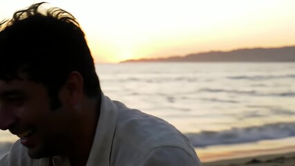 A handsome man sits on the sand laughing happily with the ocean waves and a golden sunset behind him