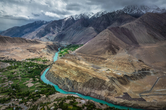 View of the turquoise river snaking through the arid landscape, contrasting with snow-capped peaks under a moody sky, Alchi Valley, Ladakh Division, India.