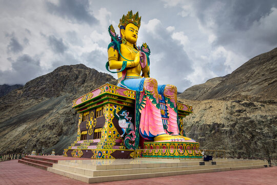 View of a golden Buddha statue sits majestically, framed by stark mountains under a brooding sky, a spiritual beacon, Diskit, Ladakh, India.