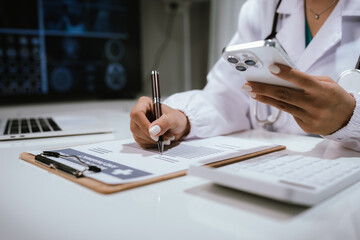 Fototapeta premium Female doctor coordinating and writing on clipboard while holding smartphone, surrounded by modern devices such as laptop, calculator and diagnostic screen in medical facility.