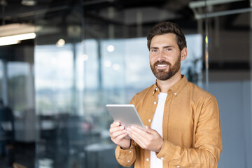 A smiling man in a modern office holds a tablet, looking directly at the camera. He has a beard and is wearing a casual shirt.