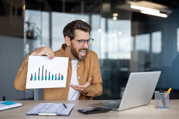 A frustrated businessman in glasses displays a declining financial graph during an online meeting, expressing dismay while sitting at his desk.