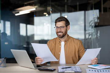 Smiling businessman in glasses reviews documents at his desk, a laptop and financial data in sight.