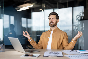 A relaxed businessman meditates at his desk in the office, hands in the mudra pose with a laptop...