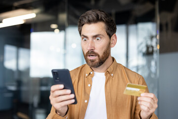 A shocked man looks at his phone screen while holding a credit card, possibly reacting to an unexpected charge or financial issue.