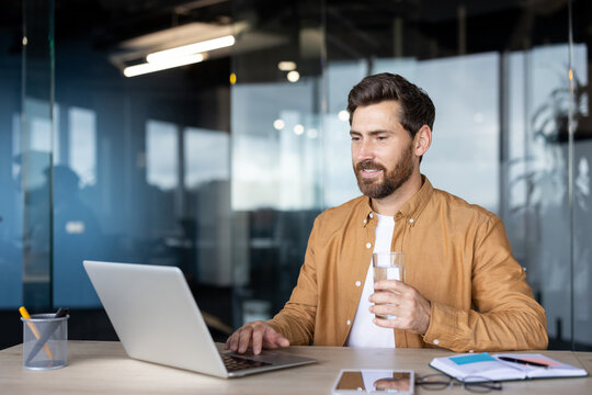 A businessman works on a laptop at his desk, smiling and holding a glass of water in a modern office setting. - Powered by Adobe