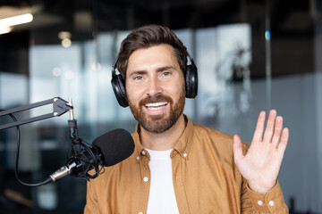 A man with a beard waves while wearing headphones and recording a podcast. He smiles near a...