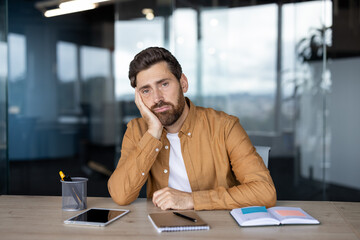 A man sits at a desk in an office, looking bored and resting his head on his hand, surrounded by...