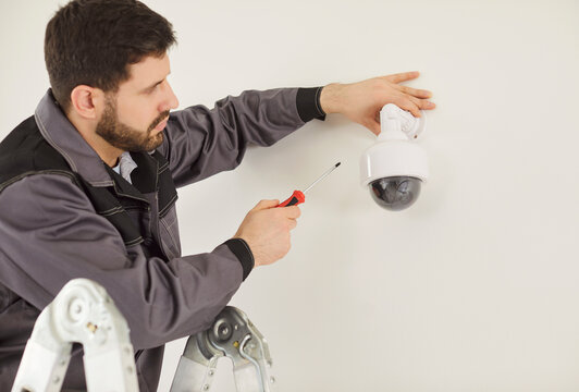 Professional technician standing on ladder and installing dome security camera on white wall. Male repairman using screwdriver focused on mounting hardware and ensuring surveillance system setup.