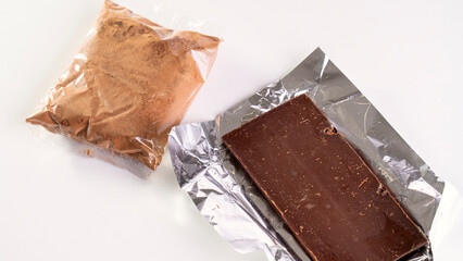 A bag of chocolate powder and a piece of chocolate are displayed on a white background. The chocolate is wrapped in foil and he is a small bar. Concept of indulgence and temptation