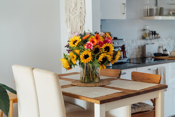 Stylish modern interior of open space white kitchen with huge multicolor summer flower bouquet in vase on wooden kitchen counter table, macrame on the wall. Cozy Design home decor. Selective focus.