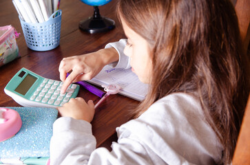 A young girl doing her maths homework using a calculator and other school supplies on a wooden table. 